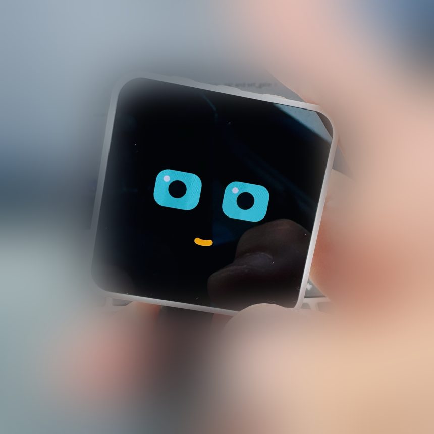 Photo of the device held in hand, showing Vibbe's code-drawn face: two teal eyes and a small orange smile on a black screen